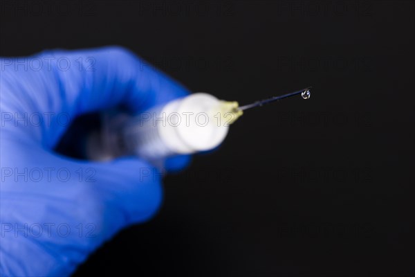 A hand with a blue medical glove is holding a syringe with a hypodermic needle and a drop of medicine