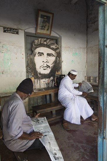 Two men reading newspapers in a room with a Che Guevara portrait