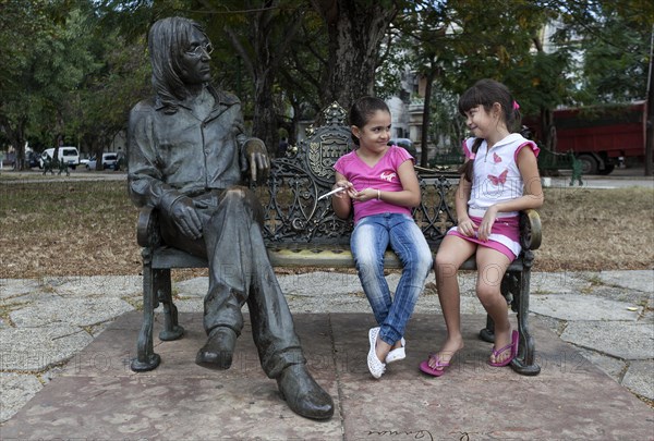 Two girls sitting on a bench next to a bronze sculpture of John Lennon