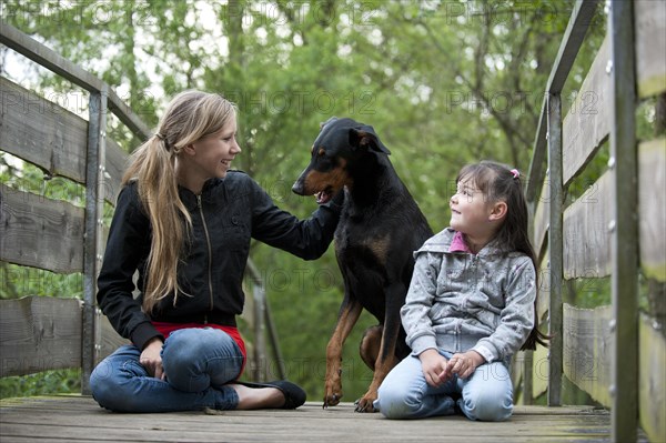 Two girls sitting with a Doberman on a bridge