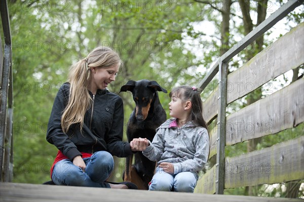 Two girls sitting with a Doberman on a bridge