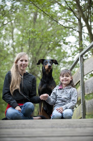 Two girls sitting with a Doberman on a bridge