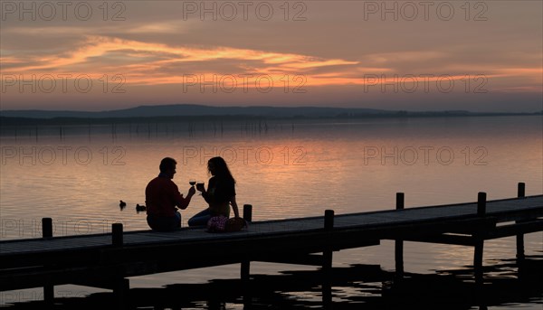 Silhouette of a couple in the twilight sitting on a wooden pier on the lake toasting with red wine glasses