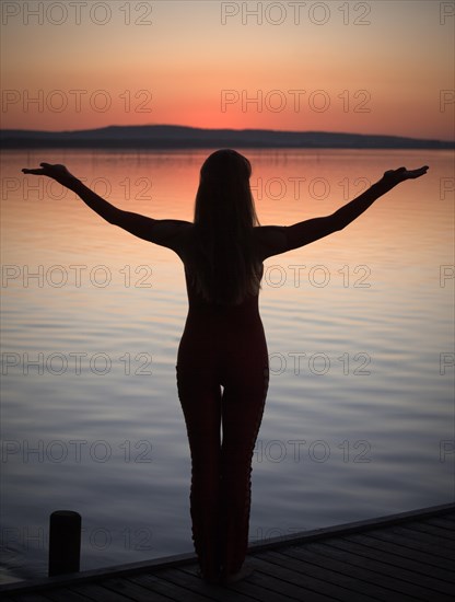 Woman with backlighting doing a yoga pose on a wooden pier at a lake after sunset