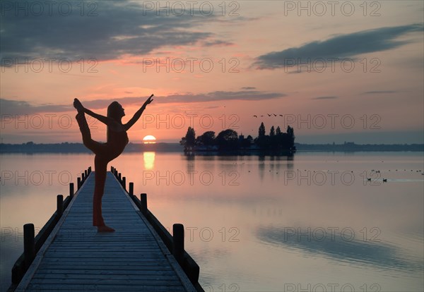 Woman with backlighting doing a yoga pose on a wooden pier at a lake in front of the setting sun