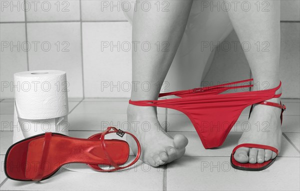 Lower leg of a young woman sitting on the toilet in black and white with red thong panties and red strap shoes