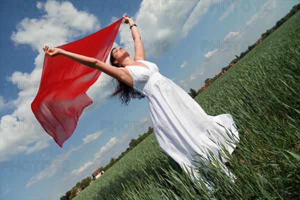 Oblique view of a young woman wearing a white summer dress in a green corn field while holding a red silk scarf fluttering in the wind