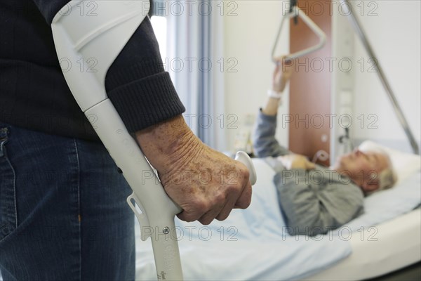 Elderly woman standing on a crutch in front of an elderly man lying in a hospital bed