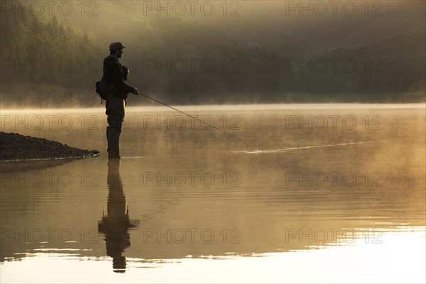 A fly fisherman standing on the shore of the Obernau reservoir at sunrise
