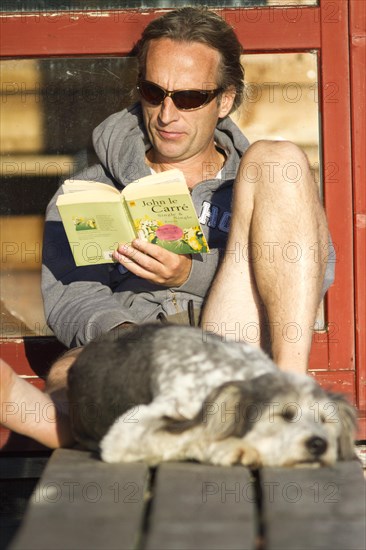Man sitting on a pier in the sun and reading a book with his dog sleeping in front of him