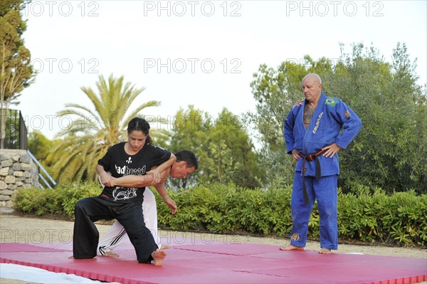 Woman and a man doing martial arts training on a sports mat in a park while being observed by their trainer