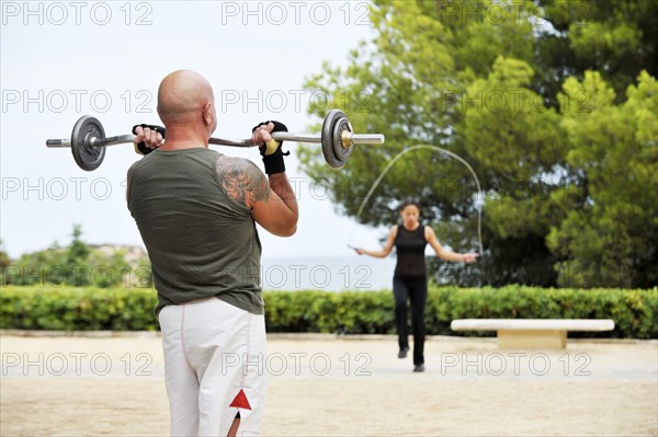 Man lifting weights and a woman jumping rope while exercising in a park