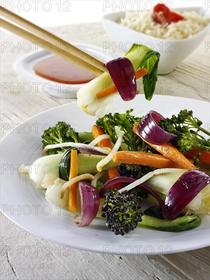 Oriental vegetarian stir fry being lifted by chop sticks with rice and a chili dipping sauce