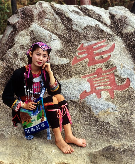 Young Miao woman wearing a traditional ethnic costume in front of rocks with a Chinese inscription