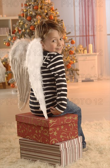 Boy wearing angel wings sitting on gifts in front of a Christmas tree