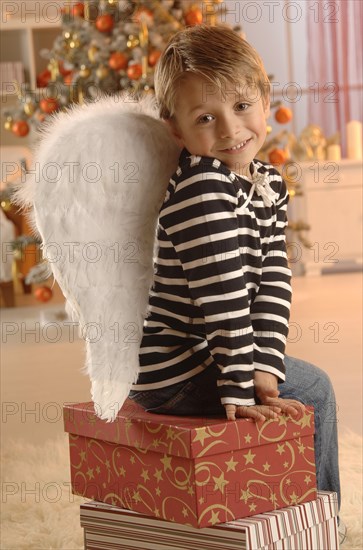 Boy wearing angel wings sitting on gifts in front of a Christmas tree