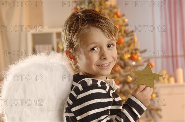 Boy wearing angel wings holding a golden star in front of a Christmas tree