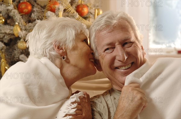 Mature couple in love wrapped in a blanket sitting in front of a Christmas tree