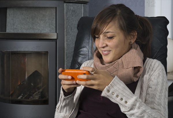 A young woman of Indonesian origin is sitting in front of a wood burner working on a smartphone