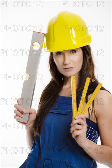 Woman wearing blue overalls and a hardhat holding a folding carpenter's ruler and a spirit level