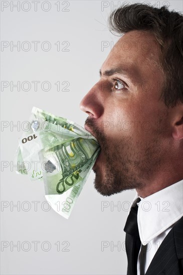 Young man wearing a suit with 100-euro notes stuffed into his mouth