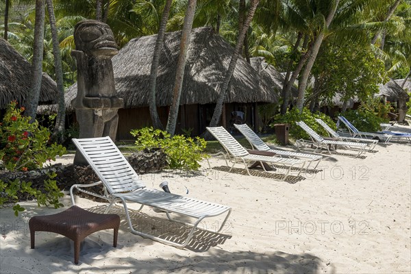 Polynesian sculpture and deckchairs in front of bungalows on the sandy beach with palm trees