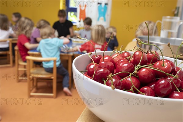 A bowl of cherries stands in the dining room in the kindergarten and children sit at the table to eat