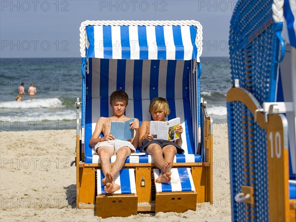 Two teenagers sit in a wicker beach chair and read a book