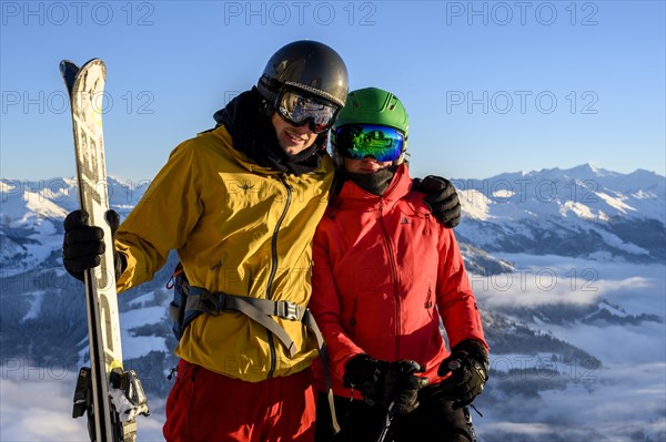 Two skiers with ski helmets and skis stand in front of a mountain panorama