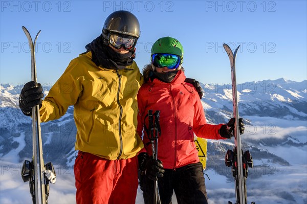 Two skiers with ski helmets and skis stand in front of a mountain panorama