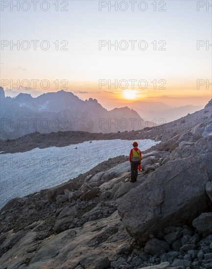 Hiker stands in rocky alpine terrain and looks at sunset over Gosaukamm with mountain peak Bischofsmuetze