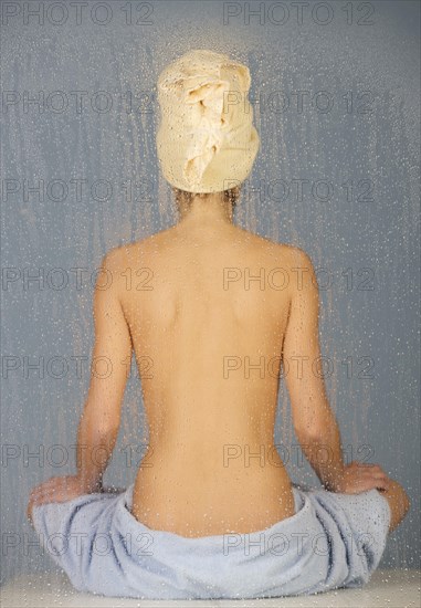 Woman sitting in a steam bath behind glass pane with drops of water, 24 years