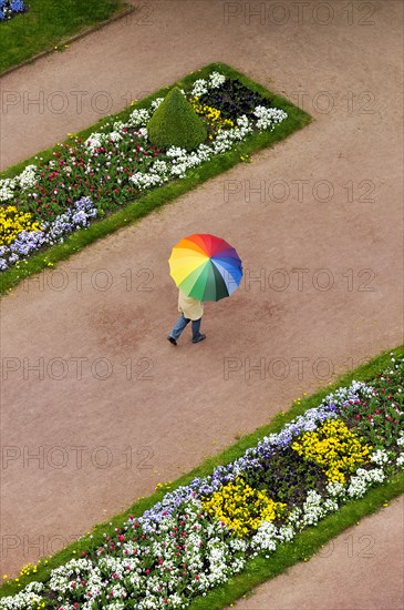 Person with an umbrella in rainbow colours on a red dirt road between flowerbeds in the Palace Gardens