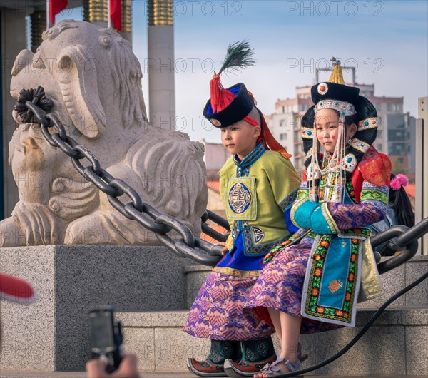 Mongolian boy in the traditional traditional traditional traditional traditional traditional traditional traditional traditional traditional costume of a man and a girl in the traditional costume of a woman sitting on a chain next to the lion statue in the central square
