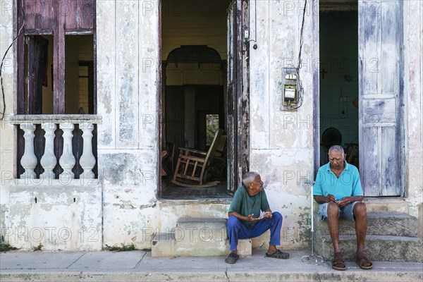 Two men having a chat at the doorsteps of their houses with decayed facades of crumbling plaster and weathered wooden doors