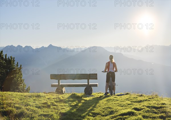 Mountain biker stands with MTB next to a wooden bench and looks into the wide light-flooded mountain landscape