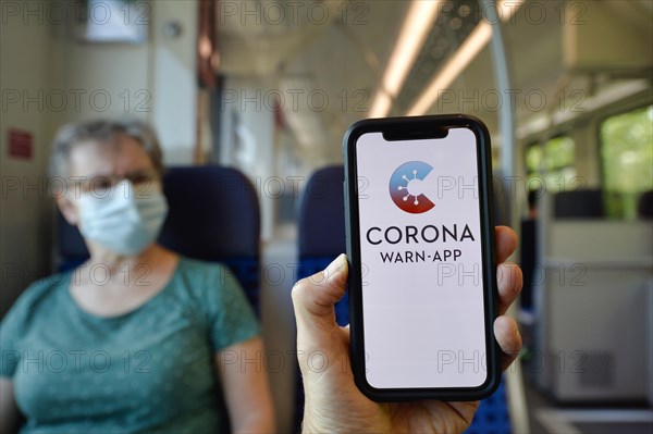 Hand holds smartphone with corona warning app in front of elderly woman with face mask sitting in S-Bahn