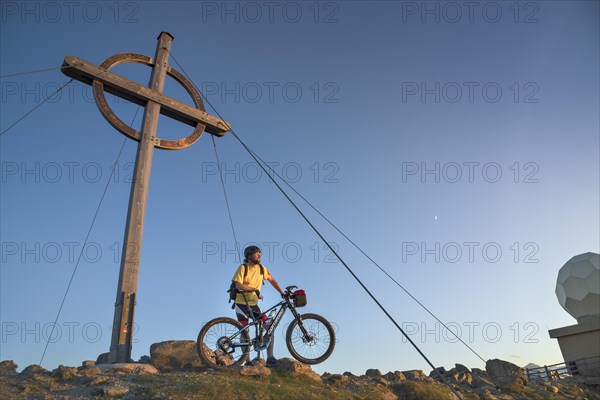 Mountain biker stands with e-MTB next to the summit cross of the Patscherkofel in the warm evening light
