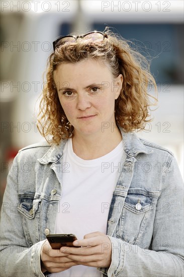 Young woman with denim jacket and sunglasses in curly hair holds smartphone in her hand