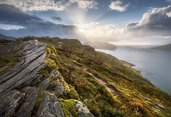 Sun breaks through dramatic clouds on the fjord with rocks and coloured moss in autumn