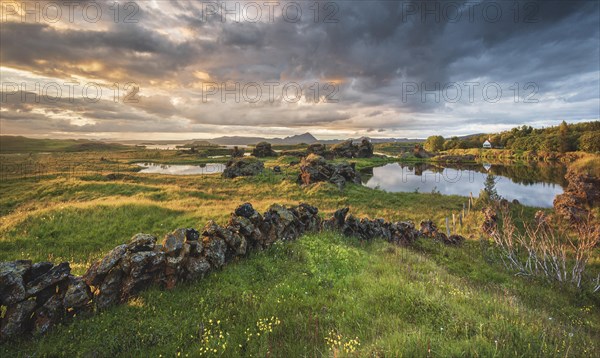 Stone wall and lava formations of volcanic rock rise from green meadow with dramatic sky in the evening light at Kalfastroend