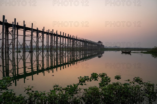 Buddhist monks walking across the U-leg bridge as the sun rises with green plants in the water
