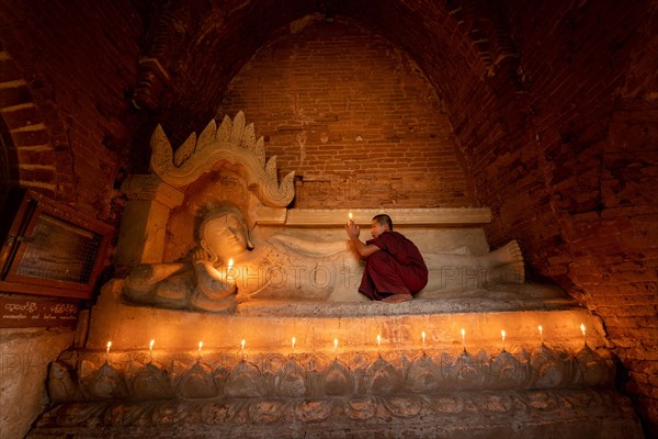 Buddhist young monk in red robe with red umbrella praying with candle in hand in a temple