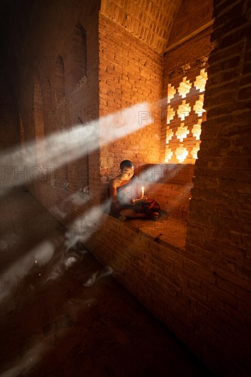 Buddhist young monk in red robe reading sitting in front of rays of light in a temple