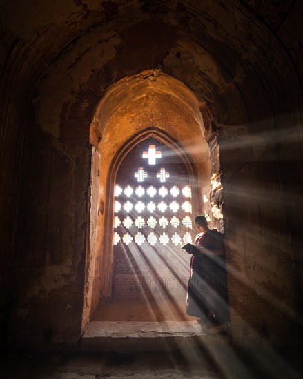 Buddhist young monk in red robe reading standing in front of rays of light in a temple