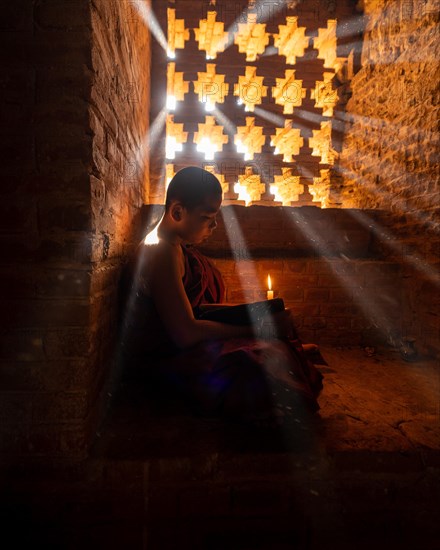 Buddhist young monk in red robe reading while sitting with a candle in front of rays of light in a temple