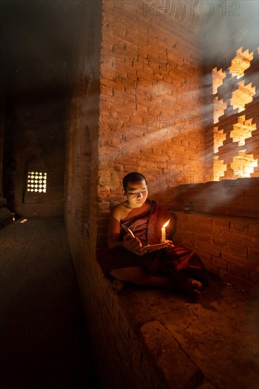 Buddhist young monk in red robe reading sitting in front of rays of light in a temple