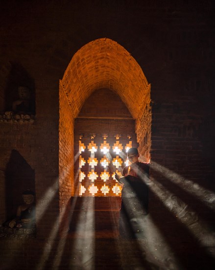 Buddhist young monk in red robe reading standing in front of rays of light in a temple