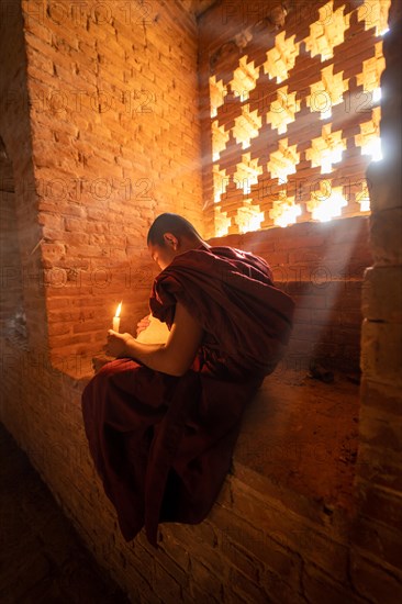 Buddhist young monk in red robe reading while sitting with a candle in front of rays of light in a temple