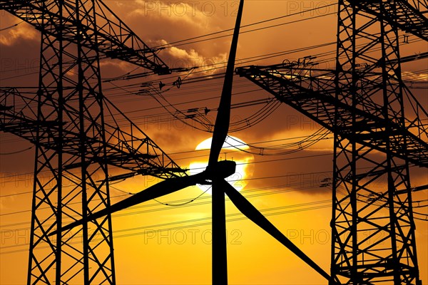 Two overhead line pylons and a wind turbine in front of the sun